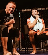 Two individuals are seated on a stage, each holding a microphone and smiling. They are in a sports-related setting with one wearing a casual black outfit and the other in sportswear, with a towel around his neck. The background includes elements of a fighting or martial arts cage.