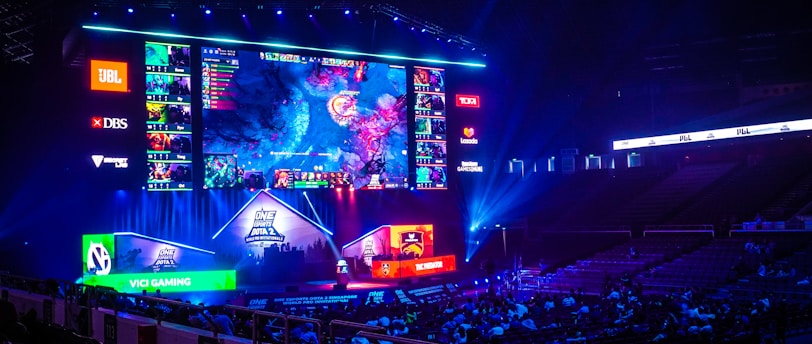 A large esports arena featuring a massive screen displaying a video game, surrounded by sponsor logos and team names. The auditorium is filled with spectators watching the gaming event. Neon blue and purple lights illuminate the stage, creating a vibrant atmosphere.