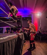 A wrestling match taking place in an indoor venue, where one participant is crouched on the top rope of the ring, while the other stands outside the ring looking at them. A crowd watches the event enthusiastically from their seats.
