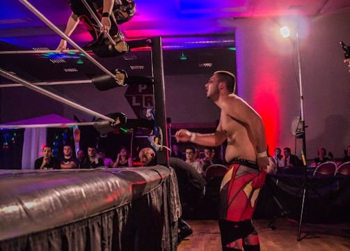 A wrestling match taking place in an indoor venue, where one participant is crouched on the top rope of the ring, while the other stands outside the ring looking at them. A crowd watches the event enthusiastically from their seats.
