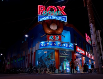 A vibrant, neon-lit facade of a gaming and sports establishment at night, featuring a large glove holding a baseball above the entrance. The building is adorned with colorful signs in both English and Japanese, with various advertisements and an illuminated sign that says 'ROX DOME'. Several people are walking towards the entrance, and bicycles are parked along the side.