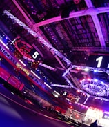 An indoor arena with vibrant purple and blue lighting, featuring large numbered screens suspended from the ceiling. The venue has a high ceiling with intricate metal truss structures and hanging lights, creating a futuristic and energetic atmosphere.