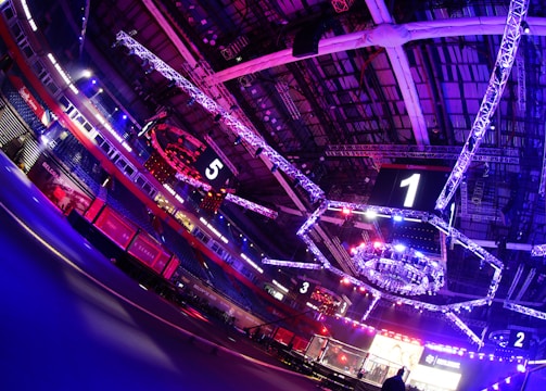 An indoor arena with vibrant purple and blue lighting, featuring large numbered screens suspended from the ceiling. The venue has a high ceiling with intricate metal truss structures and hanging lights, creating a futuristic and energetic atmosphere.