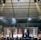 A crowded indoor arena with a cage fight taking place in the center. Audience members surround the cage, with some people involved in organizing the event inside. The arena is well-lit, and the ceiling structure is visible above.