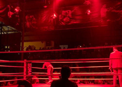 A wrestling ring is illuminated with red lighting, creating a dramatic atmosphere. Several people are visible around the ring, with silhouetted spectators in the foreground. Banners or art adorn the upper walls, and the venue has an industrial ceiling with exposed beams.