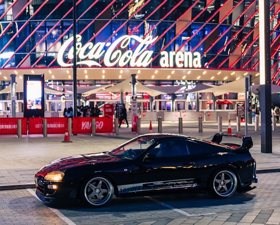A sleek black sports car is parked in front of an illuminated arena entrance, with a large sign bearing the name of a famous beverage brand. The building features a modern, geometric facade with bright red and blue lights, creating a vibrant and lively atmosphere.